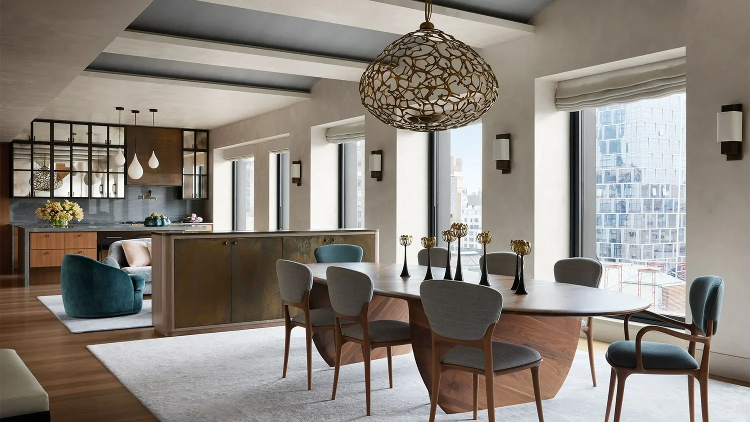 Modern dining and living area with large windows, wooden flooring, and contemporary furniture including a dining table with seven chairs, a round chandelier, a sideboard, a teal armchair, and pendant lights in the kitchen.