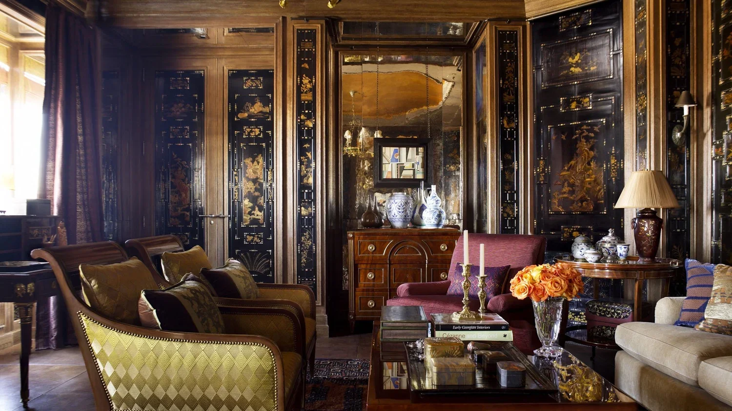 A luxurious living room with dark wood paneled walls featuring intricate gold and black Asian-style designs. The room contains a variety of upholstered sofas and chairs, decorated with patterned cushions. There is a coffee table with books and floral