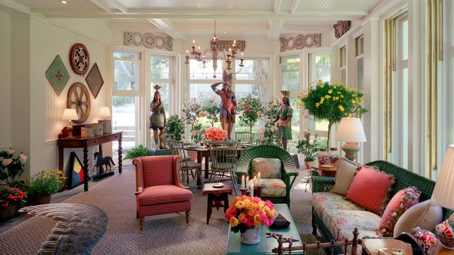 A brightly lit, traditional living room with floral decorations, vintage furniture, and statues, featuring large windows and a mix of chairs, sofas, and side tables.