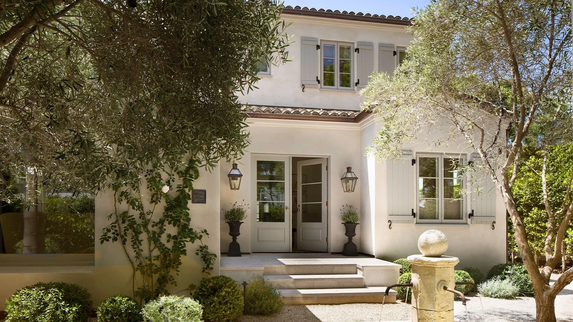 Front entrance of a white house with grey shutters, surrounded by greenery and trees, with steps leading up to a door with glass panes. There are two wall-mounted lanterns on either side of the door, and two potted plants on the steps. A decorative f