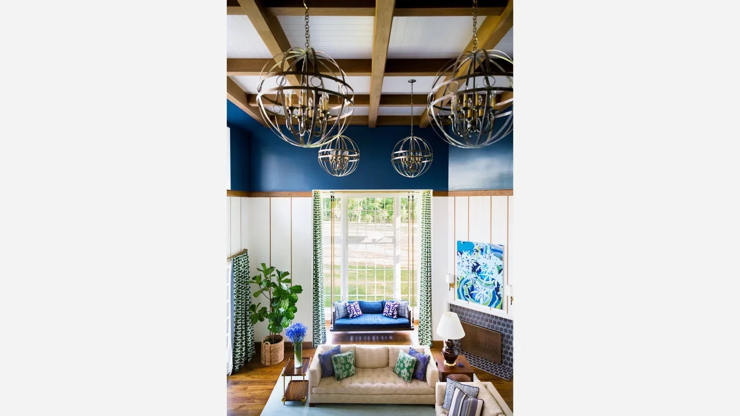 Interior of a living room with high ceiling, four spherical metal chandeliers, a large window with patterned curtains, a blue window seat, and a white sofa with decorative pillows.