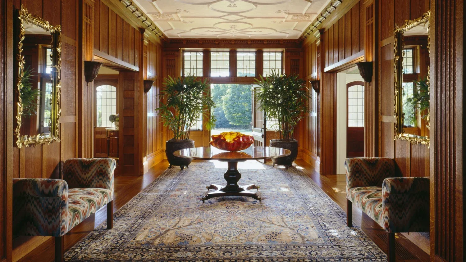 A luxurious wood-paneled room with a central round table, two potted plants, a decorative bowl, a rug, and windows showing greenery outside.