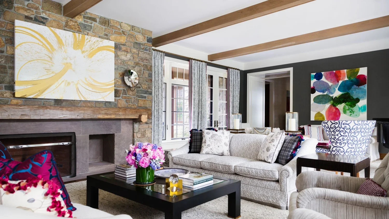 Living room with a stone fireplace, beige couch with patterned pillows, black coffee table with a vase of pink and purple flowers, dark gray accent wall with colorful abstract art, windows with gray curtains, beige armchair, and decorative candles.
