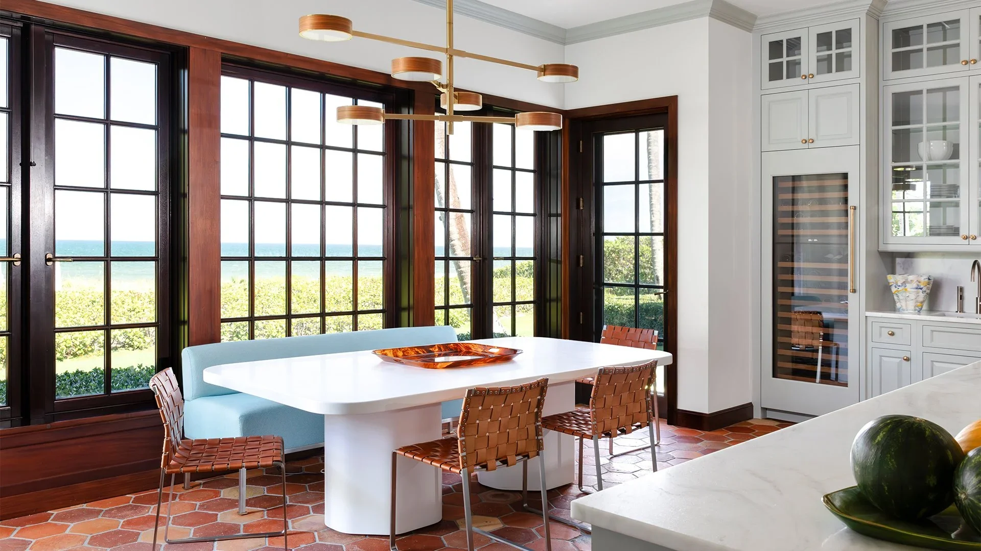 Bright dining area with large black-framed windows overlooking the ocean, white dining table, light blue banquette, woven brown chairs, and modern gold-accented chandelier, with kitchen cabinets on the right.