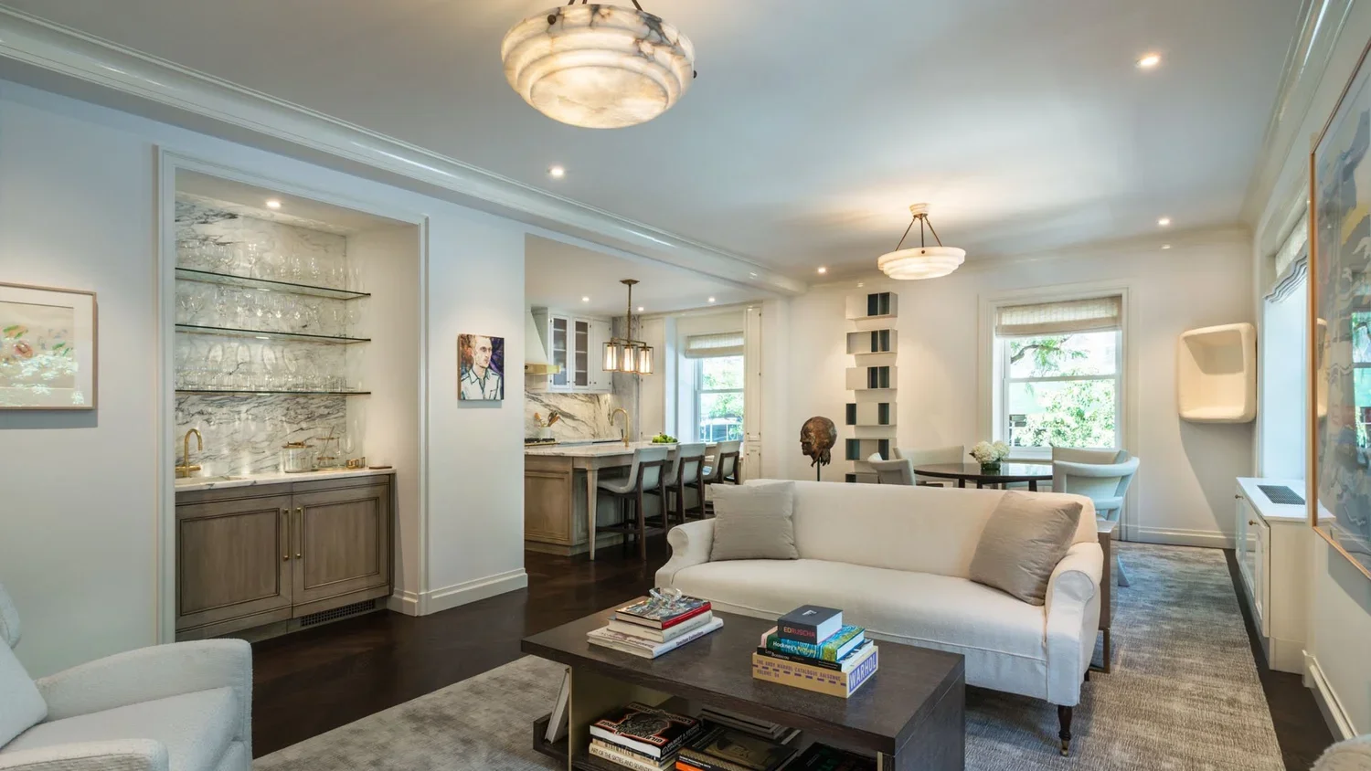 A spacious living room with white walls, hardwood floors, and modern furnishings. There is a white sofa with pillows, a dark coffee table with books, large windows with blinds, and decorative artwork on the walls. The open kitchen and dining area hav