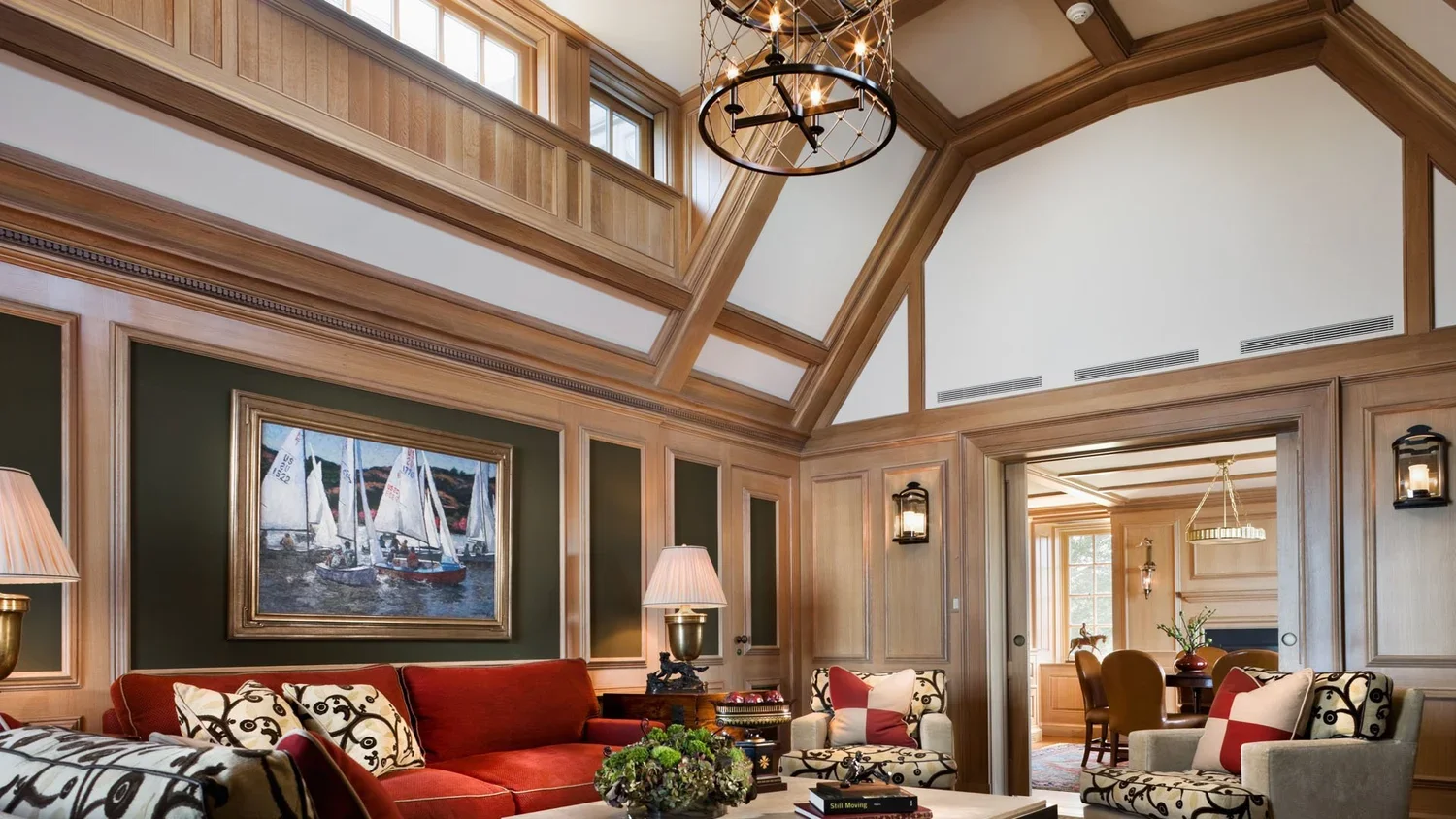 Interior view of a living room with wood-paneled walls and a vaulted ceiling, featuring a chandelier, framed painting of sailboats, a red sofa, patterned and solid accent chairs, lamps, and a doorway leading to a dining area.