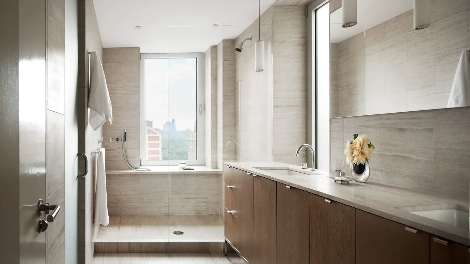 Modern bathroom with beige tile walls, a large window, a built-in bathtub, a double sink vanity with a mirror, and a vase with flowers.