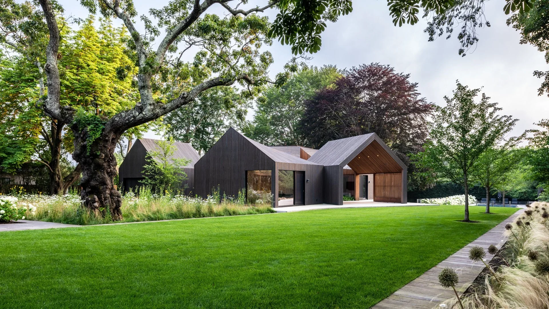 A modern house with dark wooden exterior walls and a sloped roof, situated in a lush green yard with large trees, a well-maintained lawn, and a stone pathway.