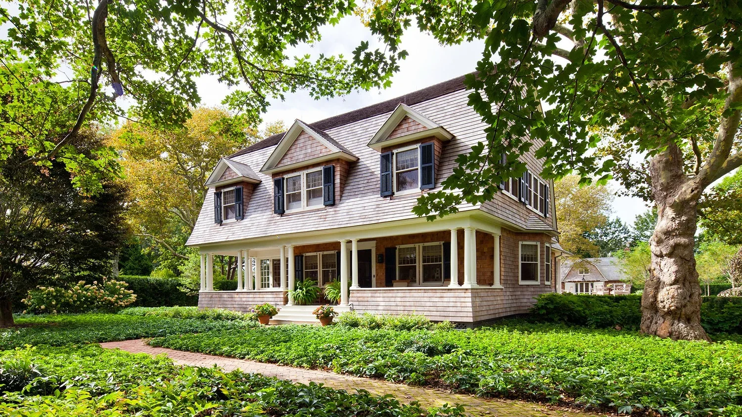A two-story house with a front porch, surrounded by lush green trees and lawn, featuring multiple windows with black shutters and a shingled exterior.