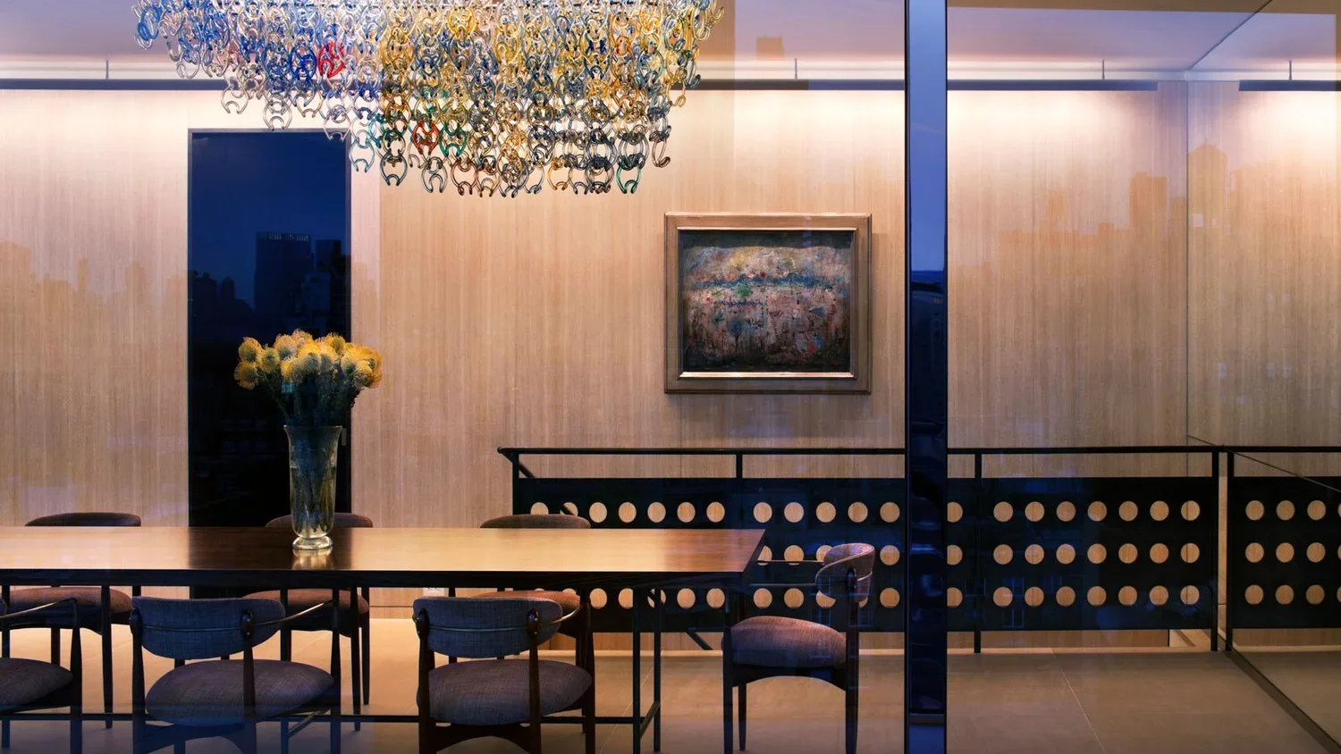 Modern dining room with a long wooden table surrounded by chairs, a vase of yellow flowers, a colorful abstract painting on the wall, and a glass chandelier hanging from the ceiling.