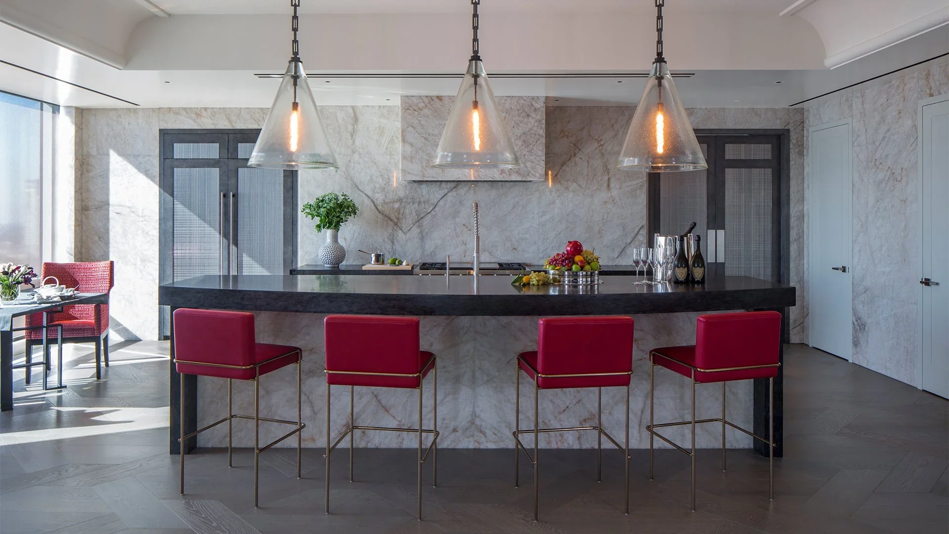Modern kitchen with black marble island, four red barstools, artful lighting, and marble backsplash.