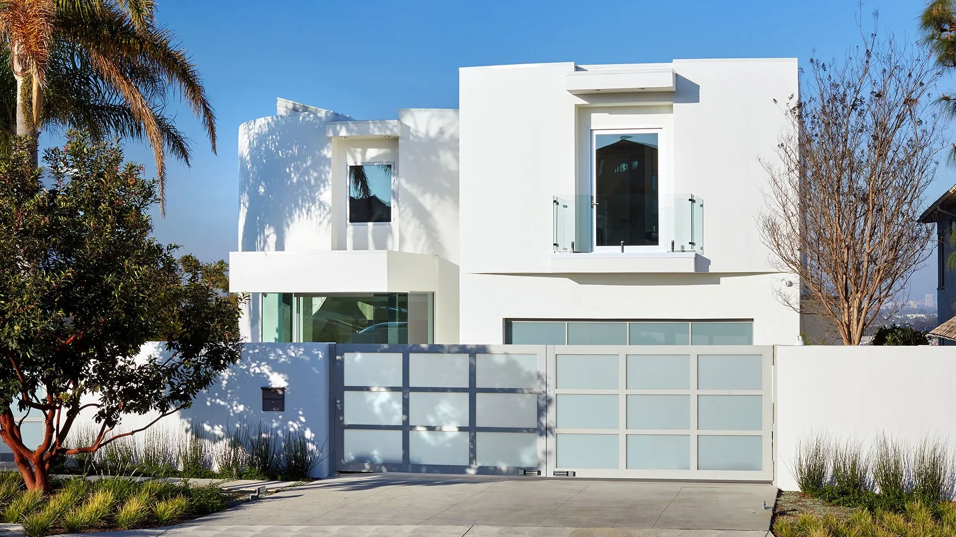 Modern white house with large windows, glass balcony, and surrounding greenery, including trees and bushes, under a clear blue sky.