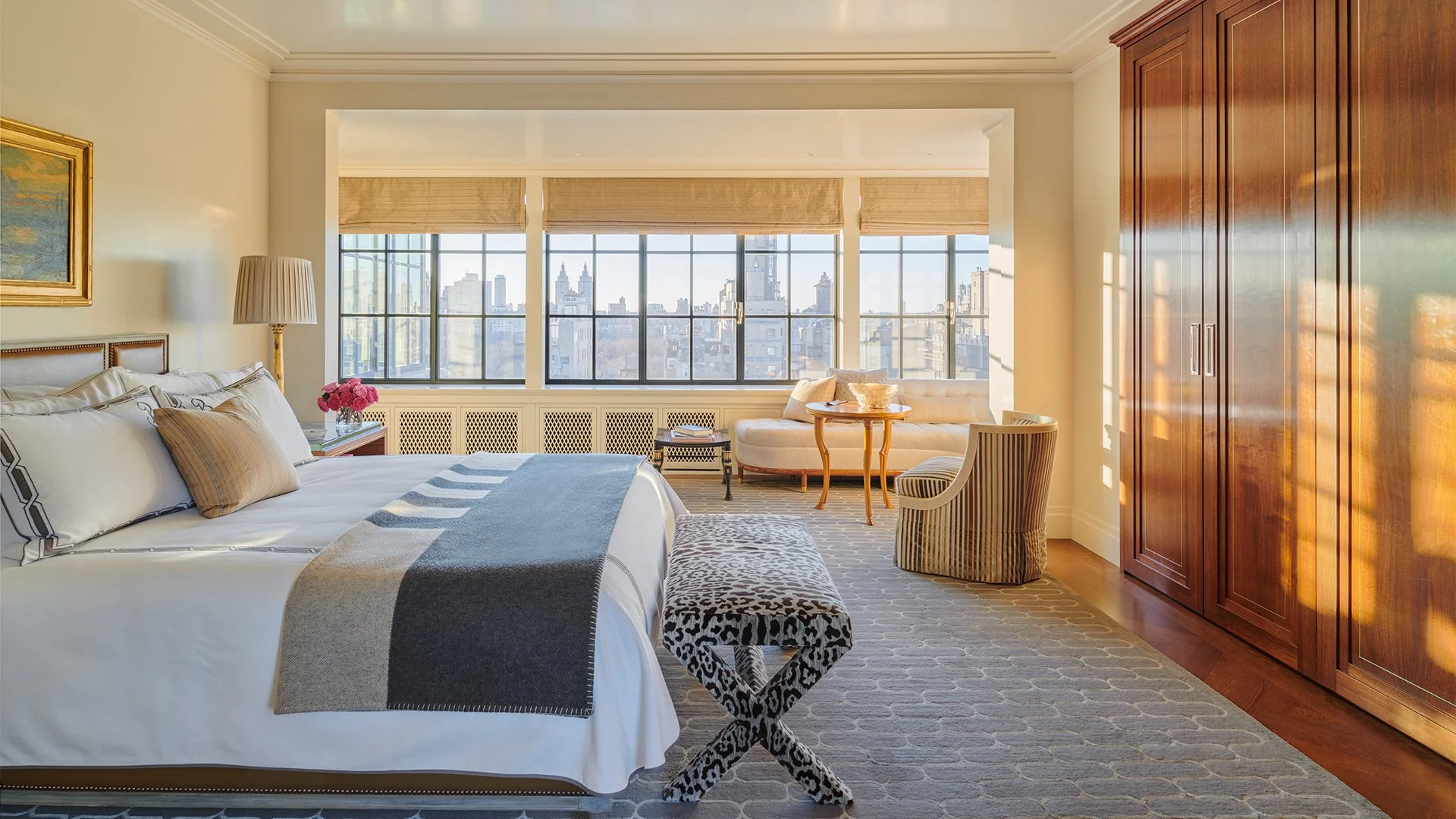 Luxurious hotel room with a large bed, a bench with animal print, a sitting area near large windows with city skyline views, and a wooden wardrobe.