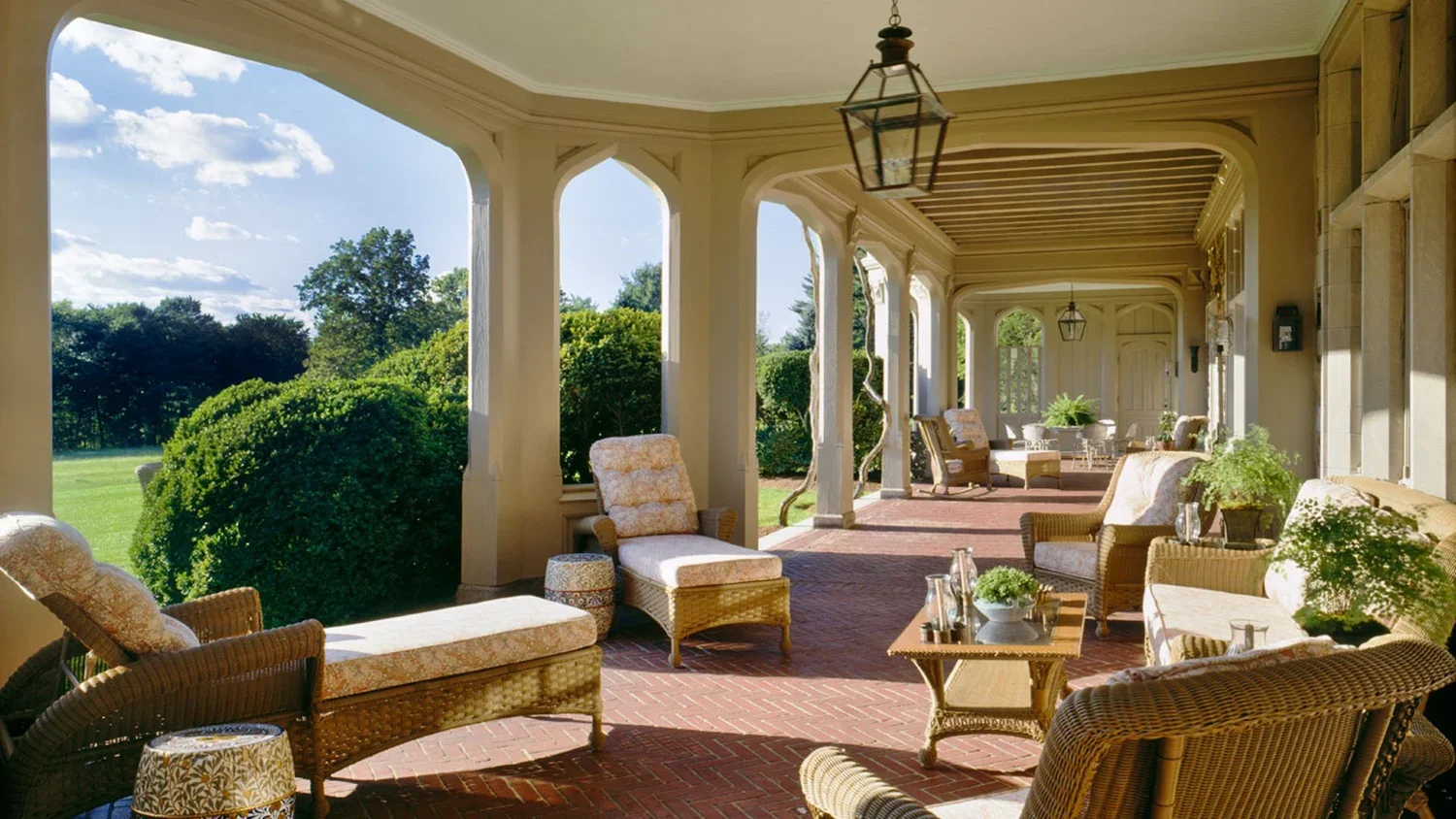 Covered porch with wicker furniture, chairs, and tables overlooking a lush green lawn and trees on a sunny day.