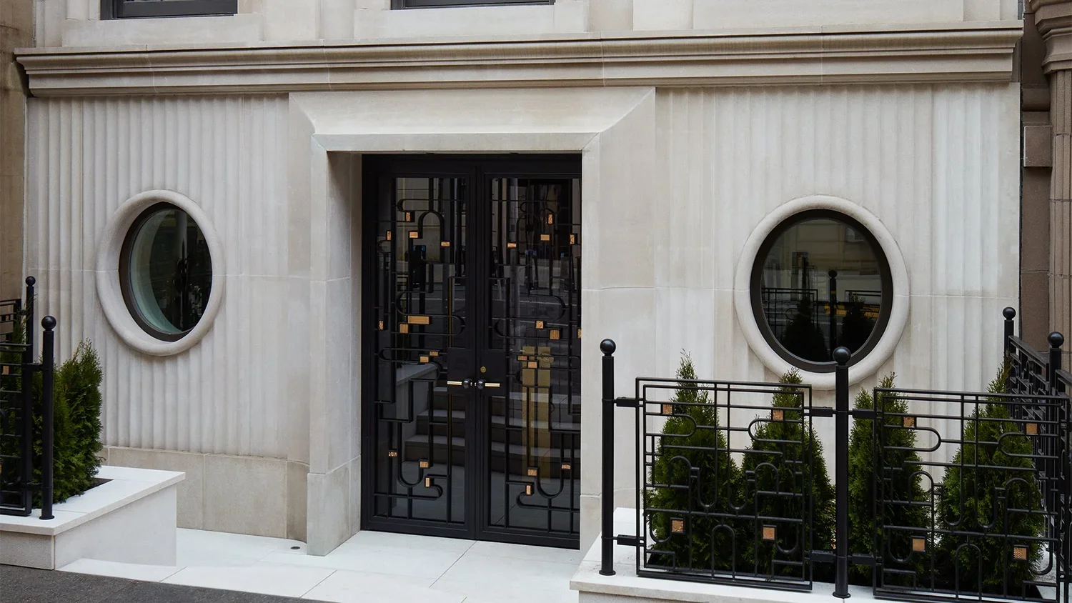 Modern building entrance with black metal gate, two round windows, and greenery in planters.