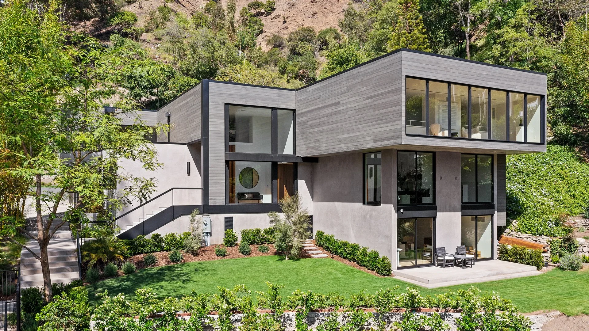 Modern multi-story house with gray exterior, large windows, and outdoor patio, surrounded by green trees and a well-manicured lawn.