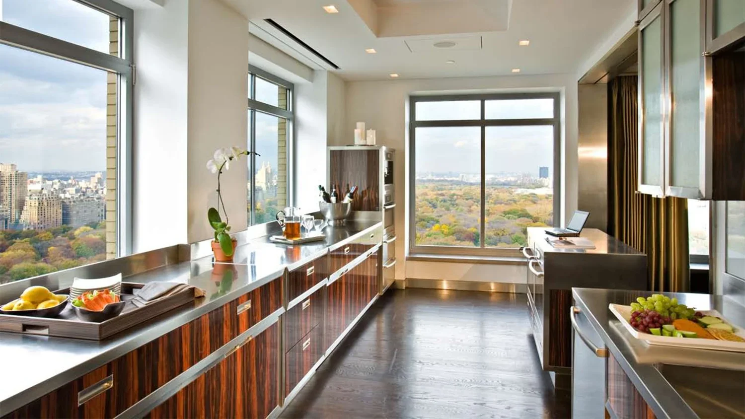Modern kitchen with large windows facing a cityscape and park, featuring wooden cabinets, stainless steel countertops, and bowls of fruit.