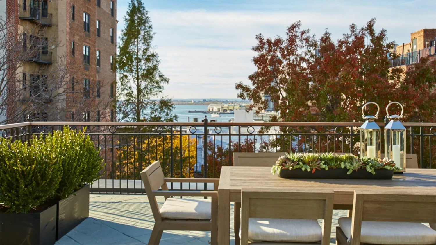Balcony view with wooden table and chairs, potted greenery, lanterns, and city skyline with water and trees in autumn.