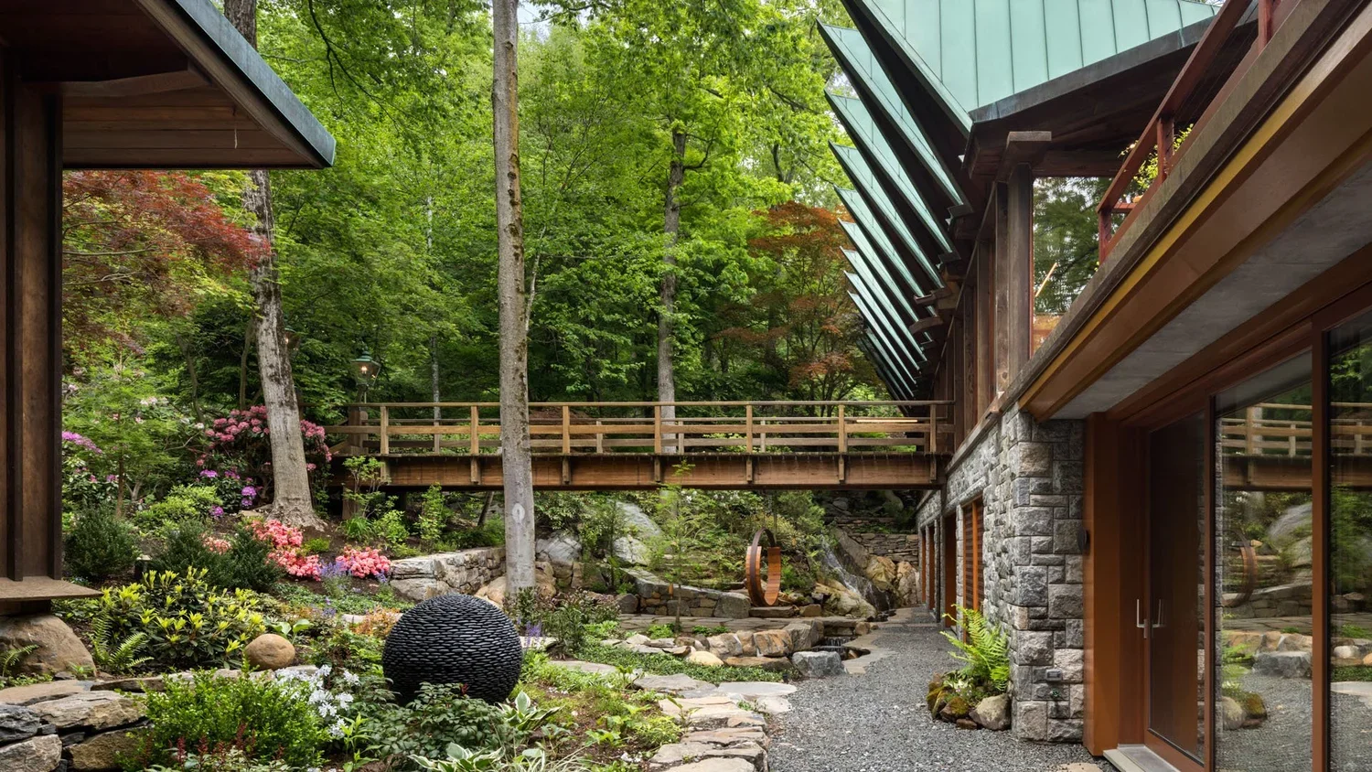 A modern house surrounded by lush green trees, with large glass windows, wooden and stone exterior, a stone pathway, and landscaped garden with colorful flowers.