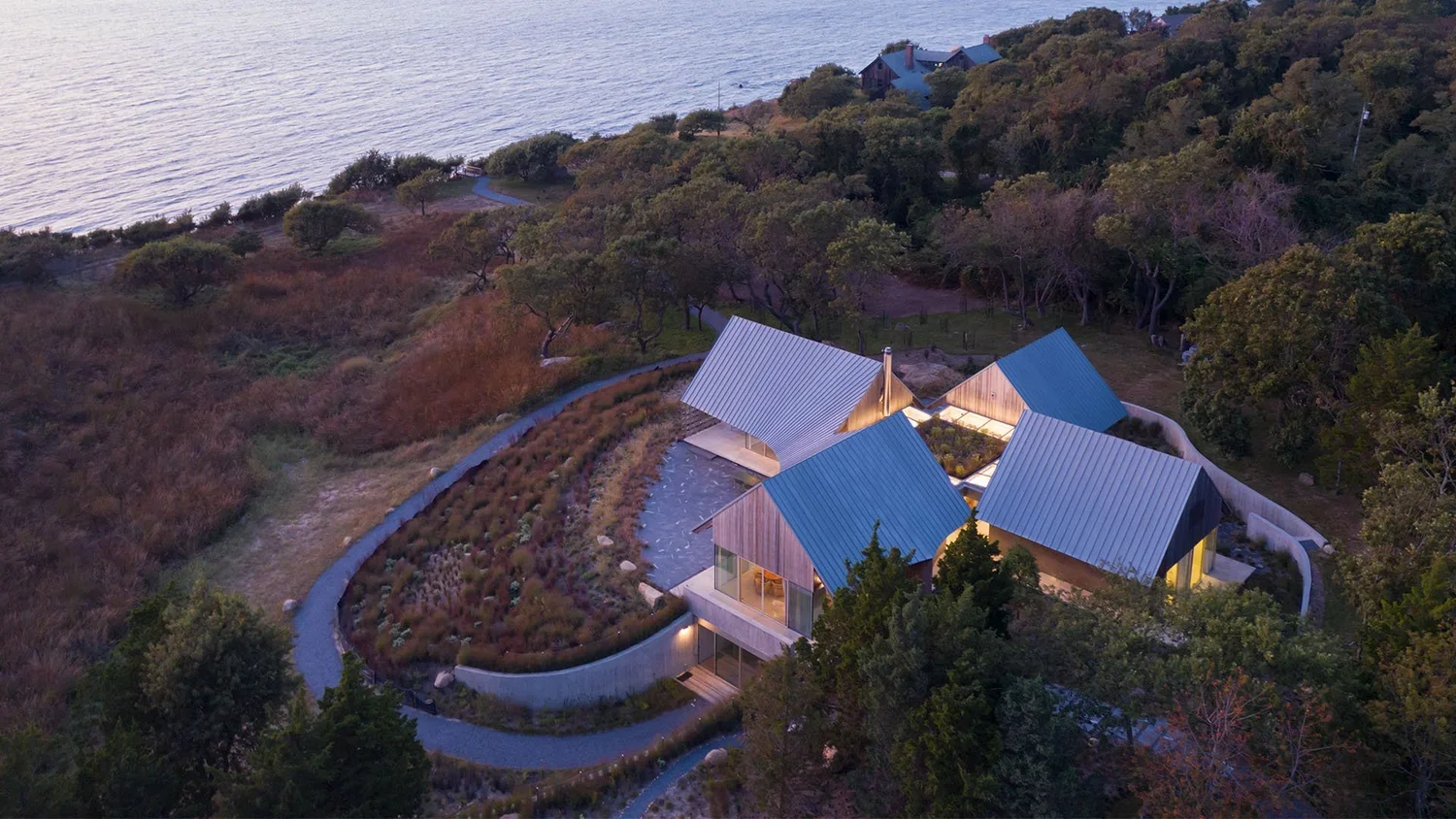 Aerial view of modern house with multiple gabled roofs, situated on a hillside near water, surrounded by trees and walking paths, with illuminated interior.