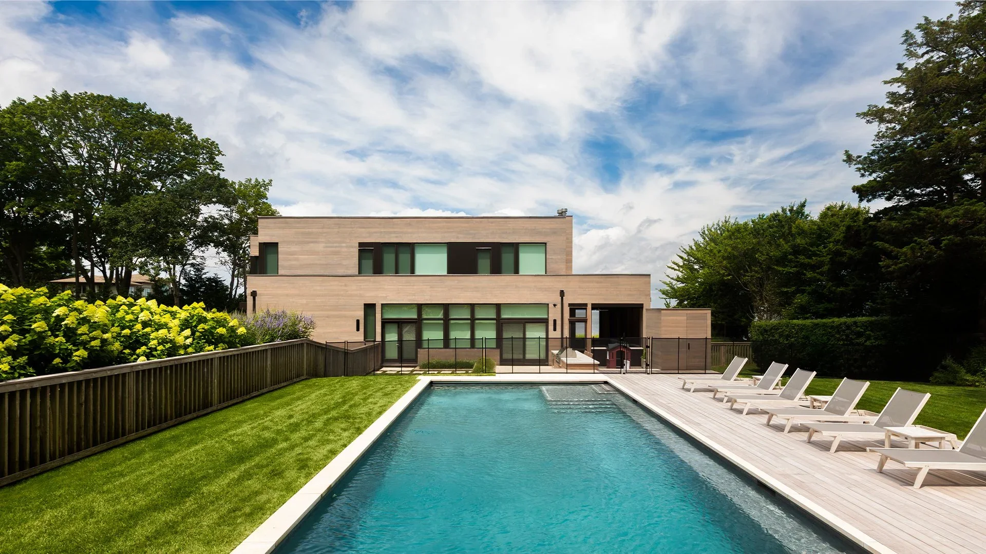 Modern house with a swimming pool in the backyard, surrounded by green grass, trees, and a wooden deck with lounge chairs, under a partly cloudy sky.