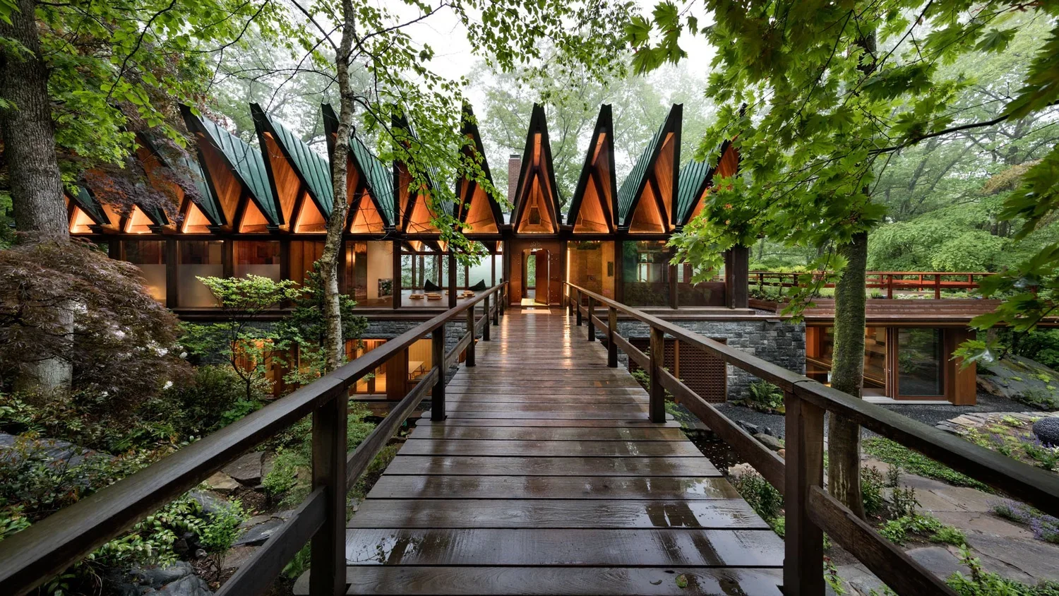 A modern house with a unique triangular roof design, situated among trees with a wet wooden walkway leading to the entrance.