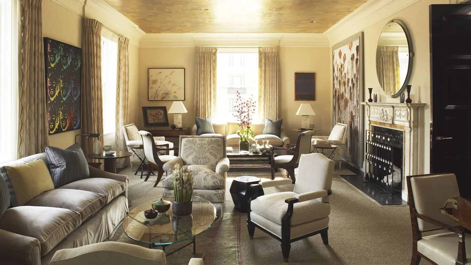 A spacious living room with a beige sofa, white armchairs, and decorative pillows, featuring a fireplace, artwork on the walls, and a large window with curtains letting in natural light.
