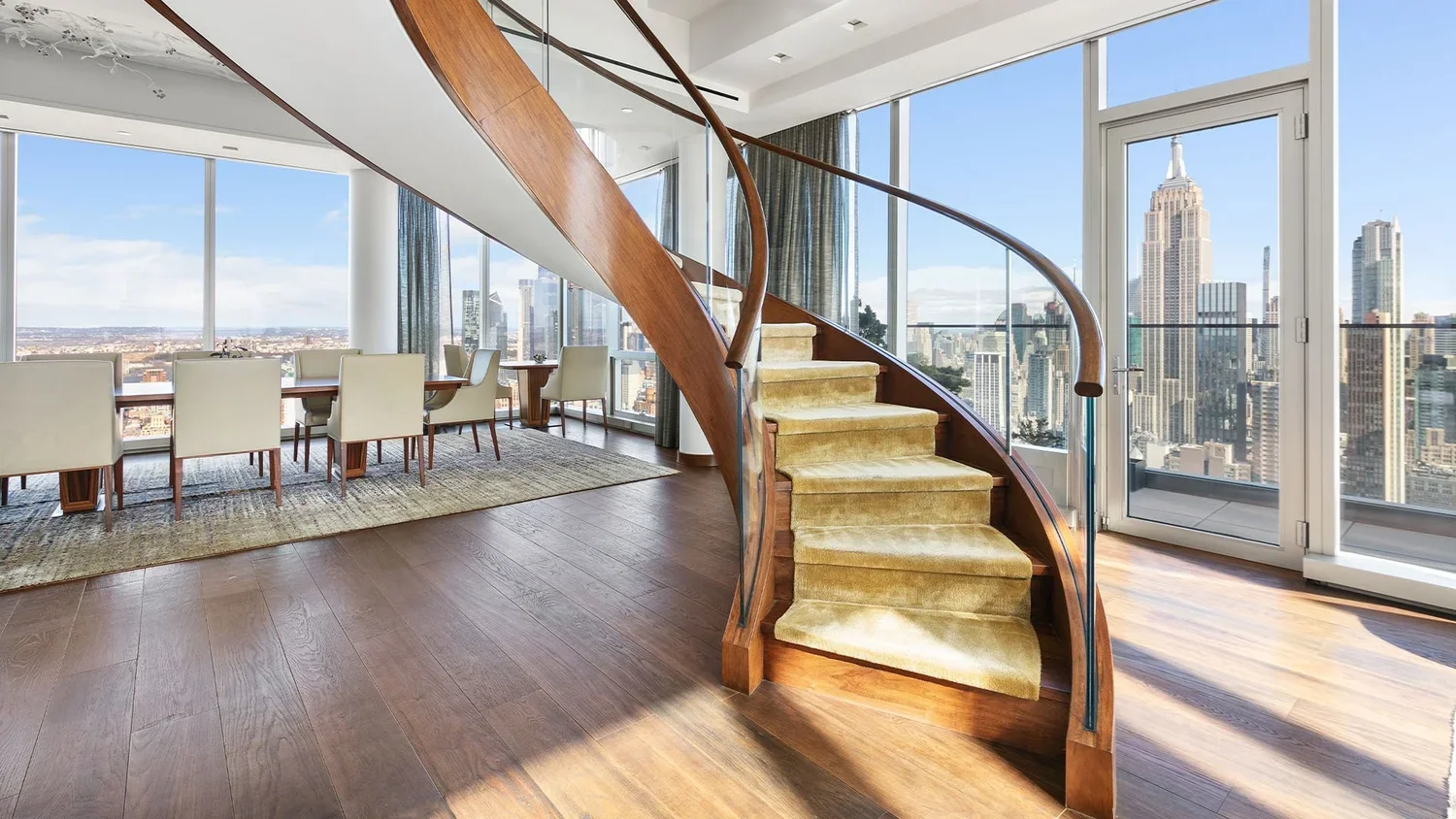 Interior view of a high-rise apartment with a curved staircase, large windows showing a city skyline, a dining table with chairs, and hardwood flooring.