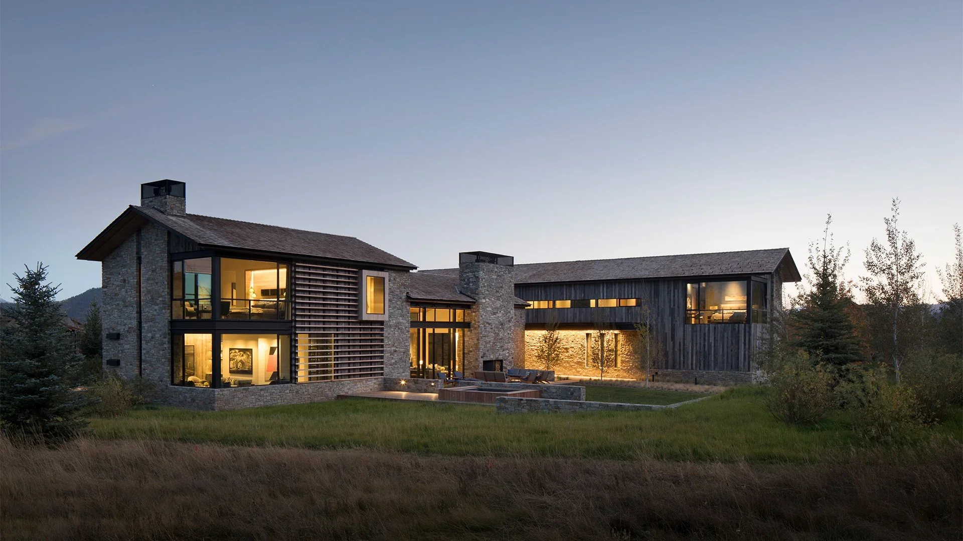 Modern house with stone and wooden exterior, large windows with warm interior lighting, surrounded by a grassy lawn and trees during dusk.