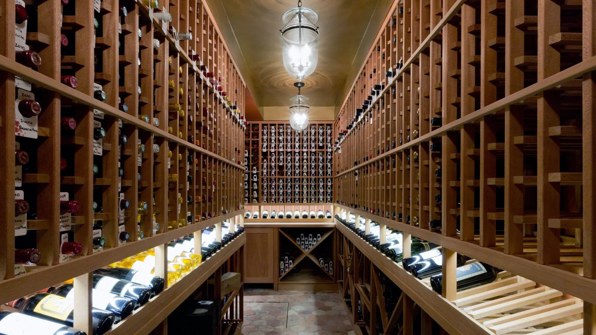 A wine cellar with wooden racks holding bottles of wine, illuminated by overhead lights and a chandelier, with a stone floor.