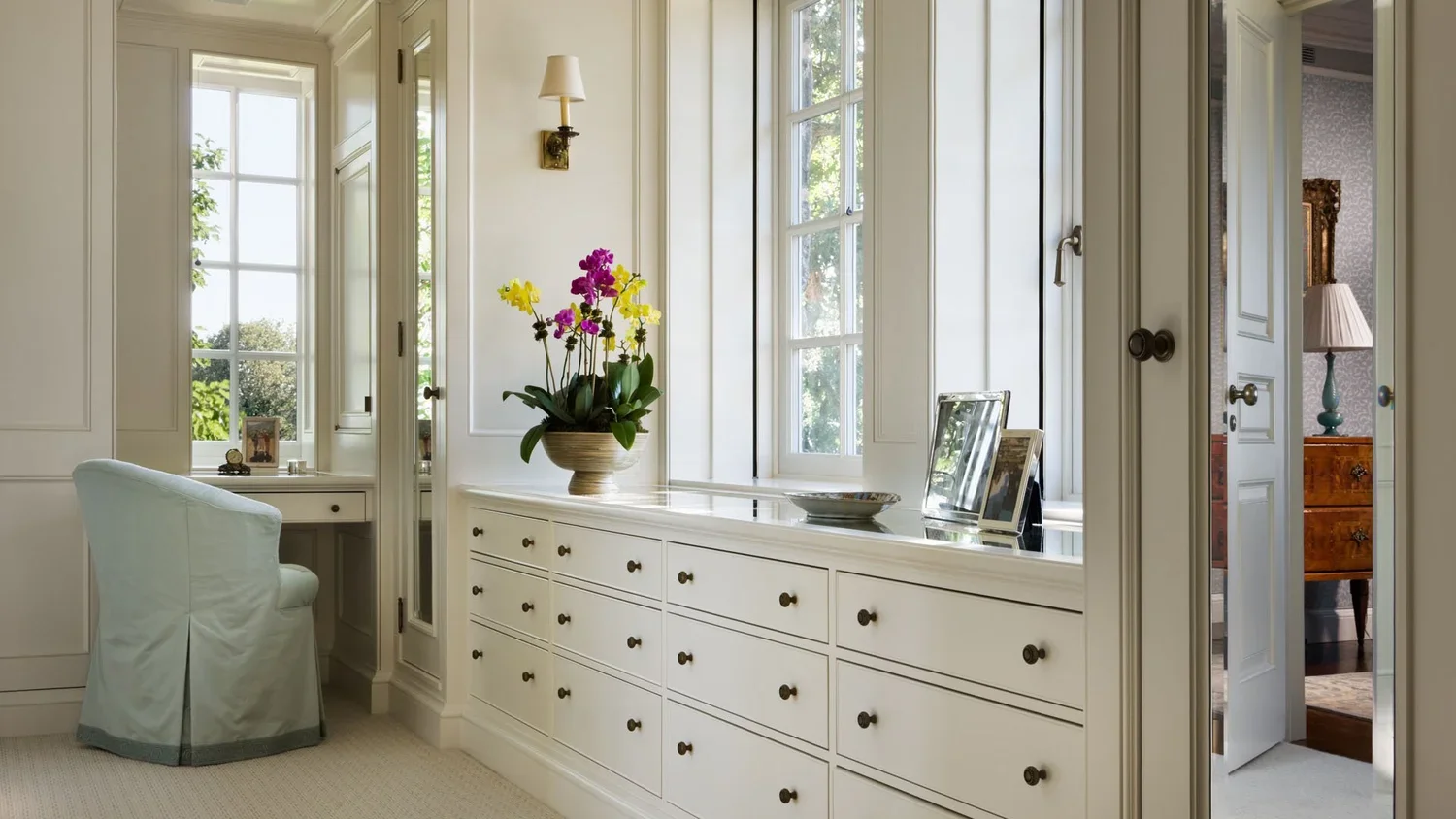 Interior of a bright, white room with large windows, a white dresser with small black knobs, a vase of colorful orchids, framed photos, and a light blue armchair.
