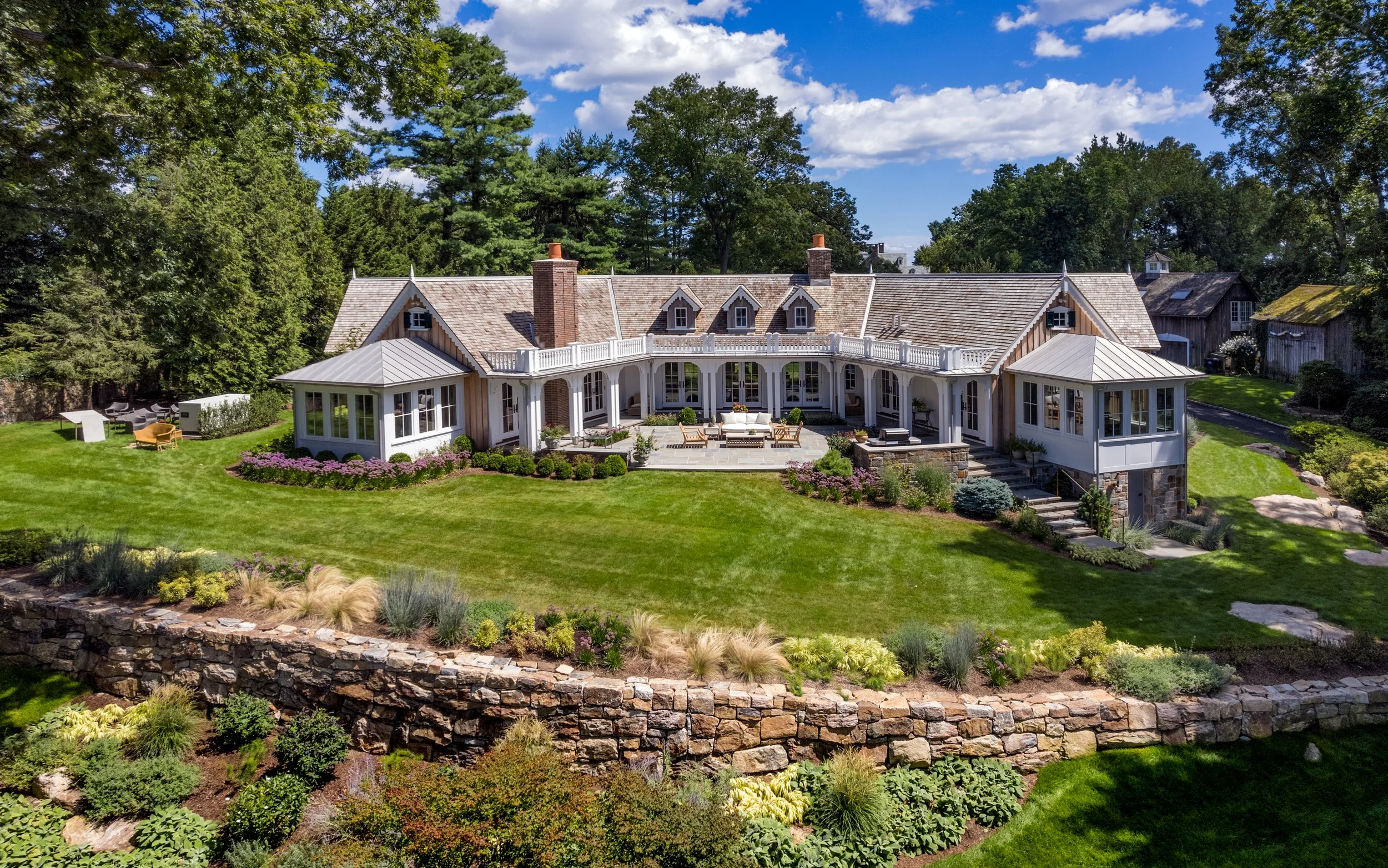 Large house with manicured lawn, flower beds, and stone retaining wall under a partly cloudy blue sky