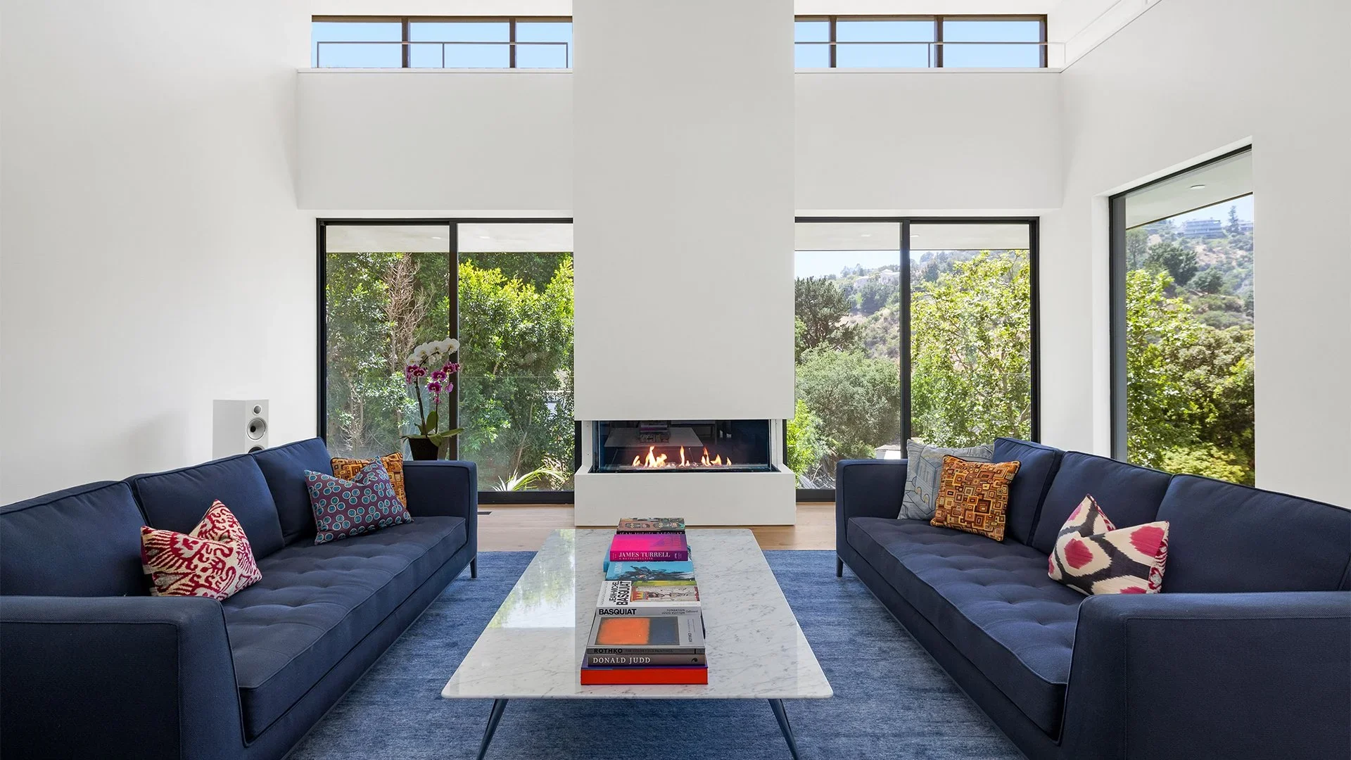 Modern living room with two navy blue sofas facing each other, each with colorful patterned pillows. A white marble coffee table with books is between the sofas. A sleek, white fireplace is centrally located on the back wall, with large windows on ei
