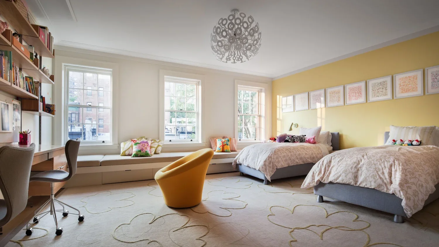 Bright bedroom with two beds, large window seat, built-in bookshelf, study desk, yellow accent wall, colorful pillows, and a unique circular chandelier.