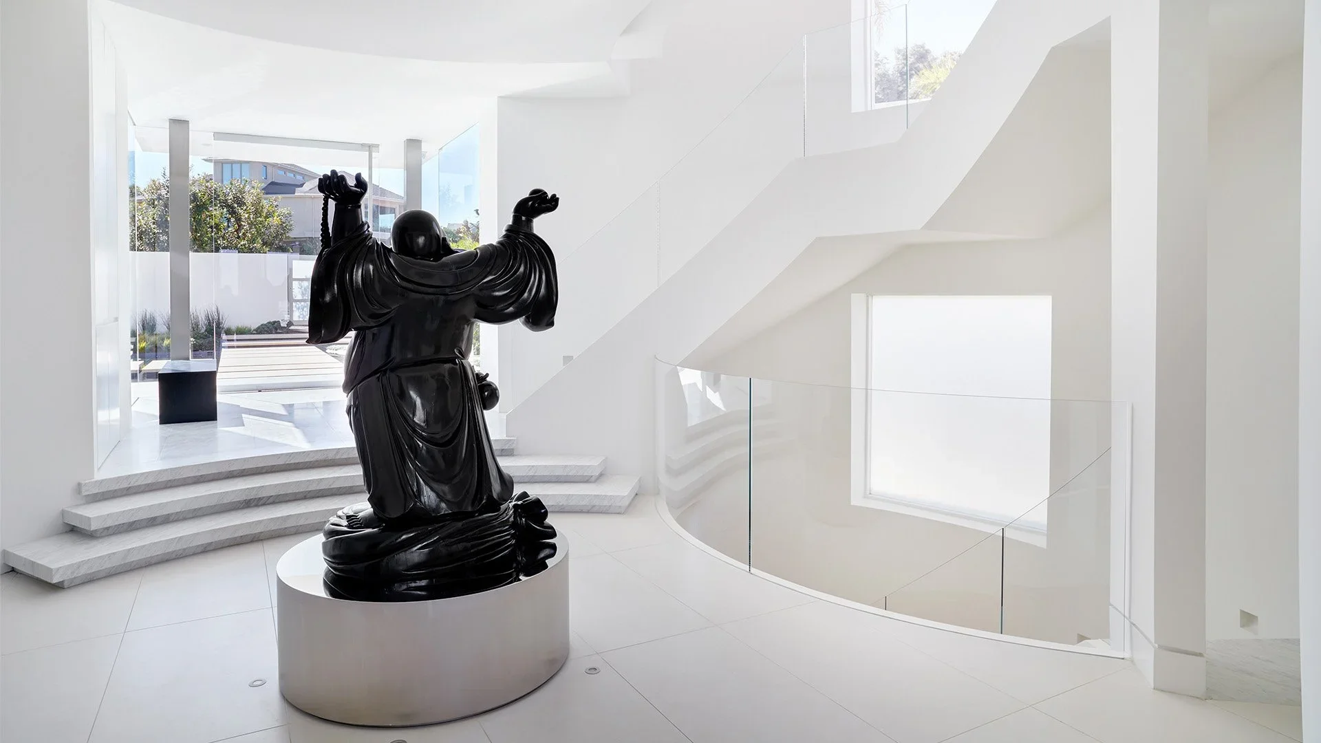 Interior view of a modern white building with a black sculpture of a person in a robe, standing on a round white pedestal near stairs and large windows.