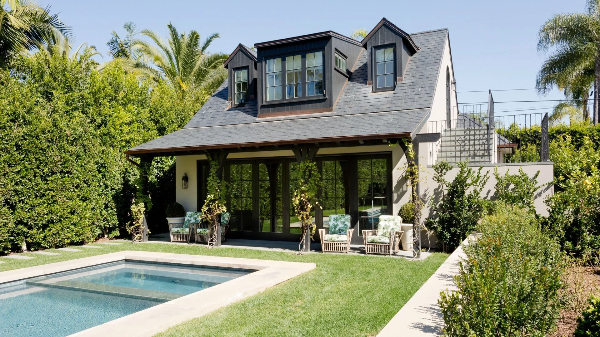 A backyard view of a modern house with a swimming pool, outdoor seating, and lush greenery including palm trees and hedges under a clear blue sky.