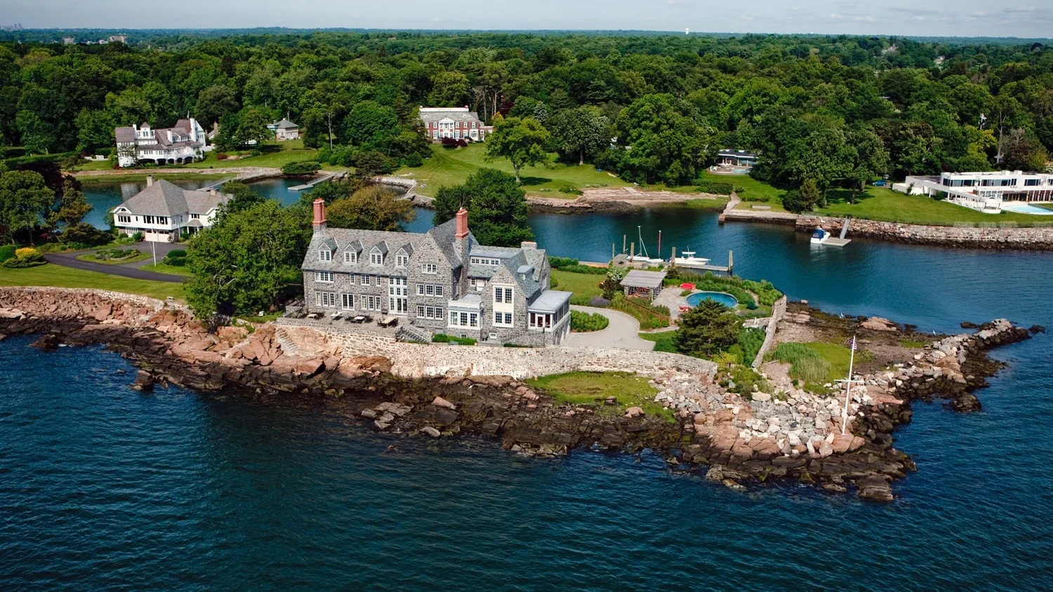 Aerial view of a large stone house on a rocky coastline with a small pier, surrounded by lush green trees and other waterfront homes.