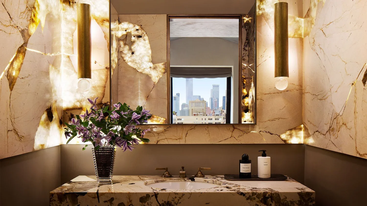 Luxurious bathroom with marble walls and sink, large mirror, decorative lighting, and a city skyline view through the window. Vase with purple flowers and bathroom essentials on the countertop.