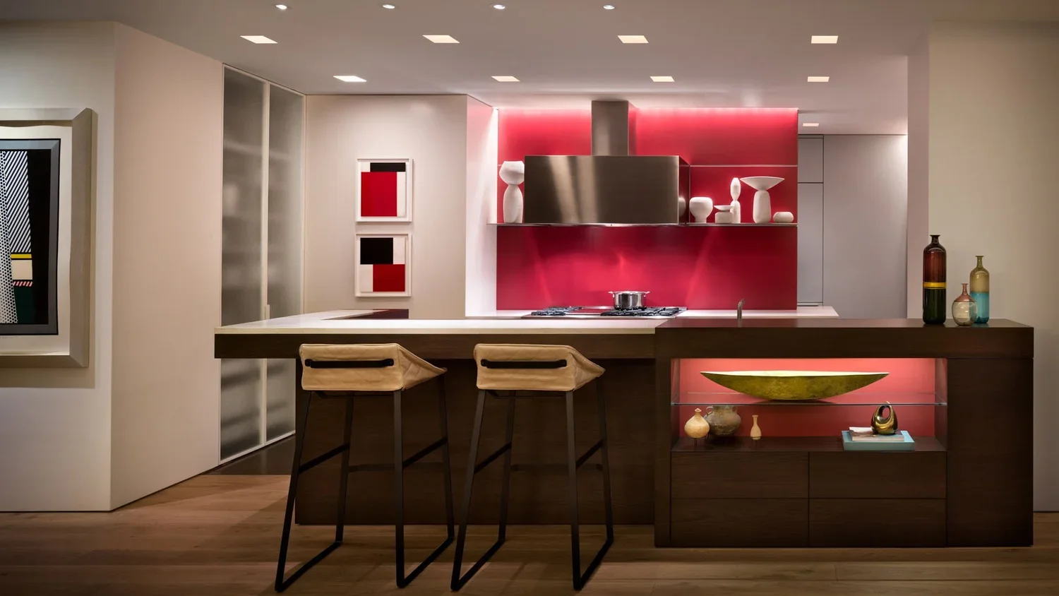 Modern kitchen with dark wood island, beige chairs, red backsplash, white sculptures, and colorful vases on a side cabinet.