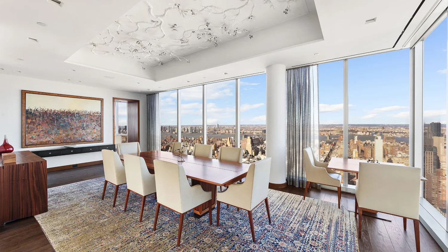 Modern dining room with large windows showing city skyline, six cream-colored chairs around wooden dining table, abstract painting on white wall, wooden sideboard with red decorative vase, patterned area rug, ceiling adorned with white floral sculptu