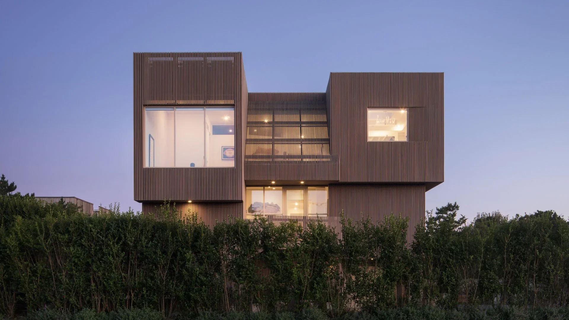 Modern multi-story house with wood siding exterior, large windows, and a flat roof, surrounded by trees at dusk.