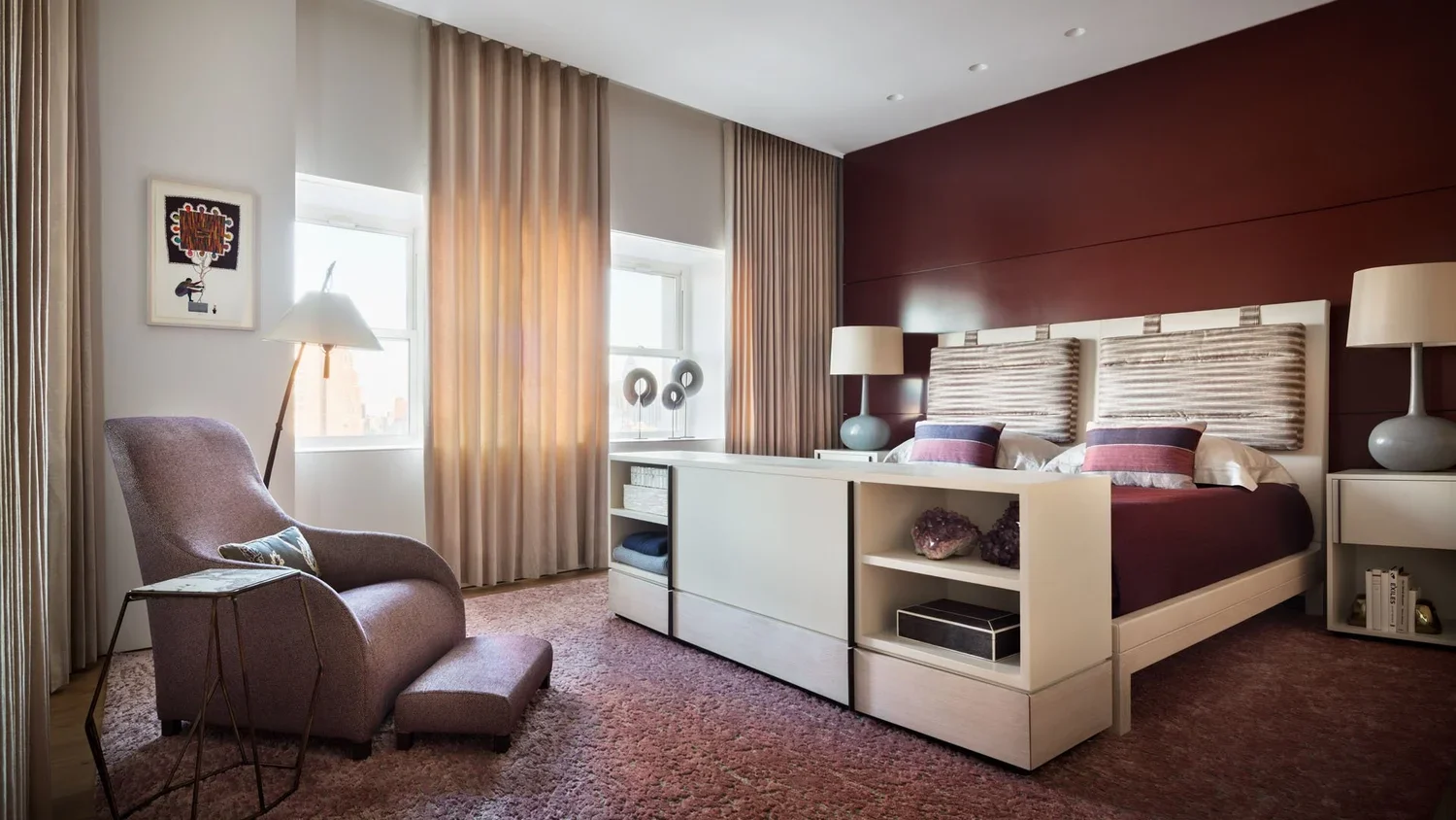 A modern bedroom with a white bed, purple and beige accents, two nightstands with lamps, a pink armchair with a footstool, and beige curtains near windows.