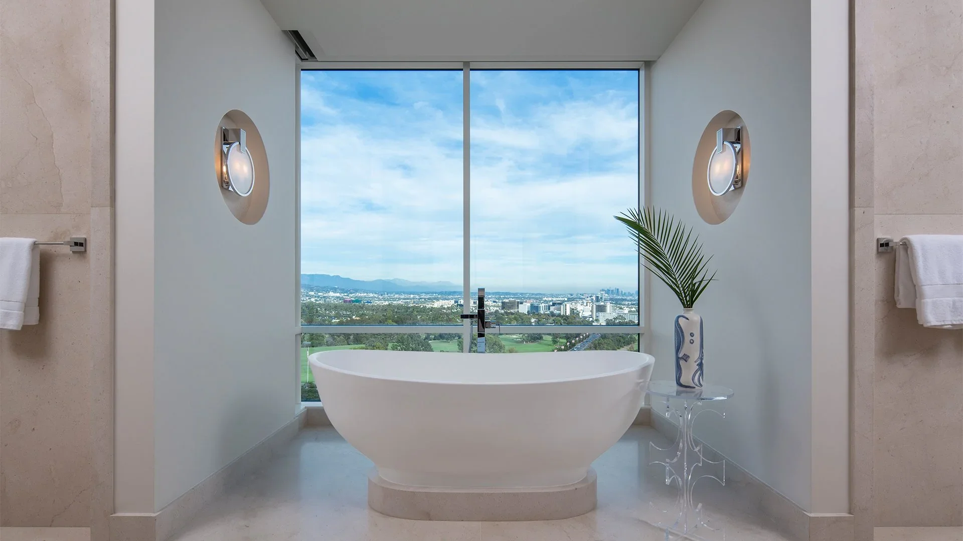 Modern bathroom with a freestanding bathtub in front of a large window showing a cityscape and blue sky.