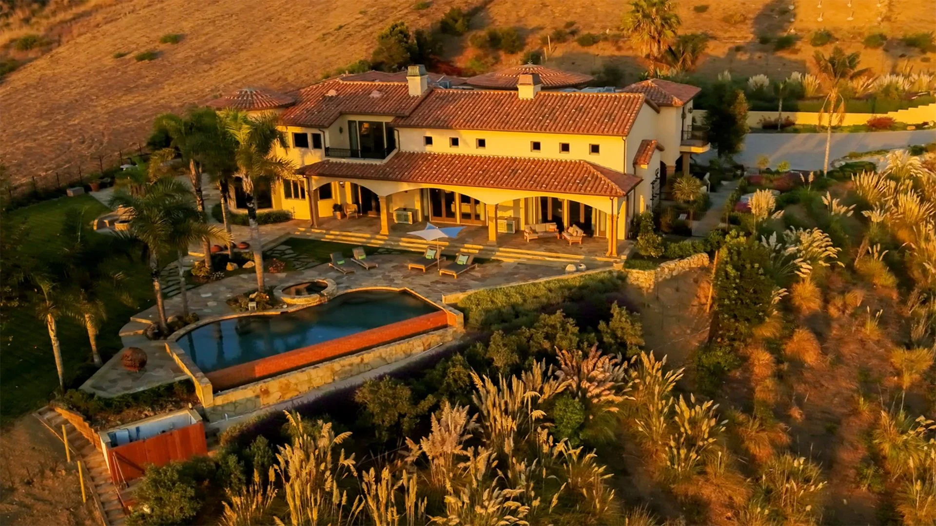 An aerial view of a large mansion with a terracotta roof, surrounded by a landscaped yard with palm trees, a swimming pool, outdoor seating, and a fire pit, set against a hillside during sunset