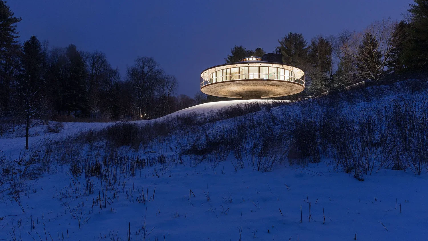 A round, modern house with large glass windows illuminated at night, situated on a snow-covered hill surrounded by trees.