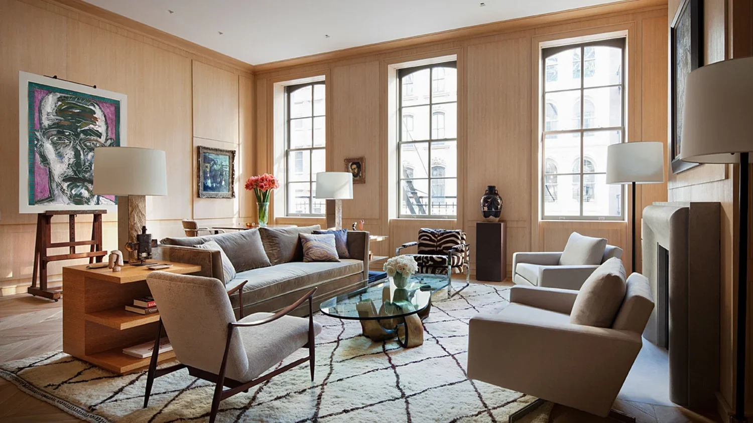 Living room with wooden panel walls, large windows, a beige sofa, white armchairs, a glass coffee table, and various artwork and decorative items.