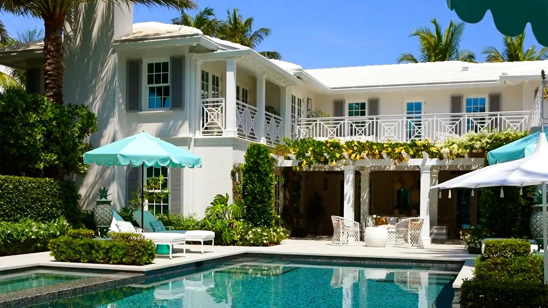 A luxurious white house with a swimming pool in the backyard, shaded lounge chairs with umbrellas, and surrounded by lush greenery and palm trees, under a clear blue sky.