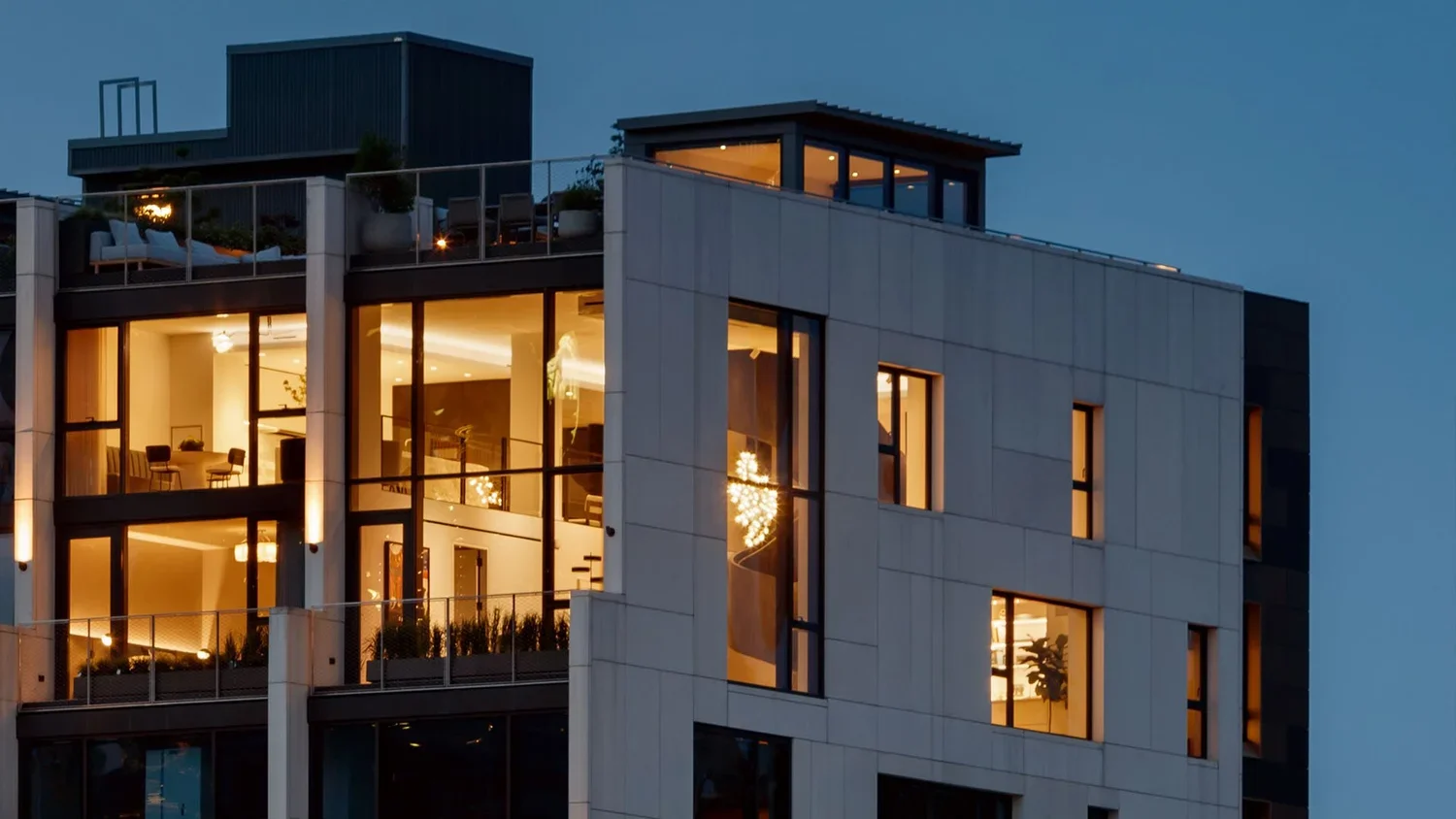 Modern multi-story apartment building with large glass windows illuminated from within at dusk.