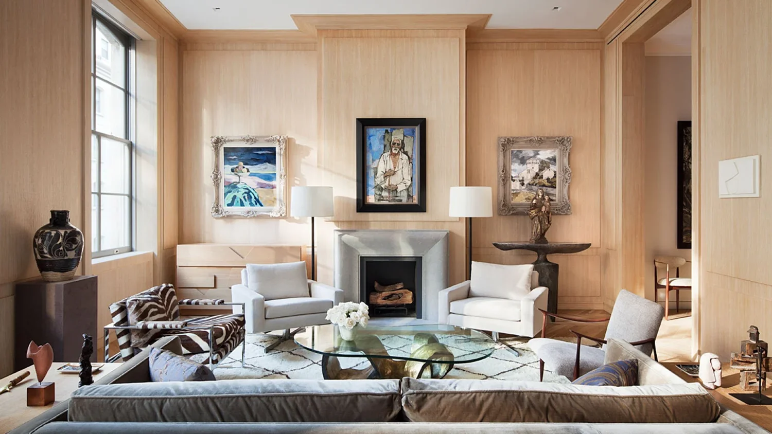 A living room with beige wooden walls, two white armchairs, a zebra-patterned armchair, a glass coffee table with decorative objects, and artworks hanging on the wall above a fireplace.
