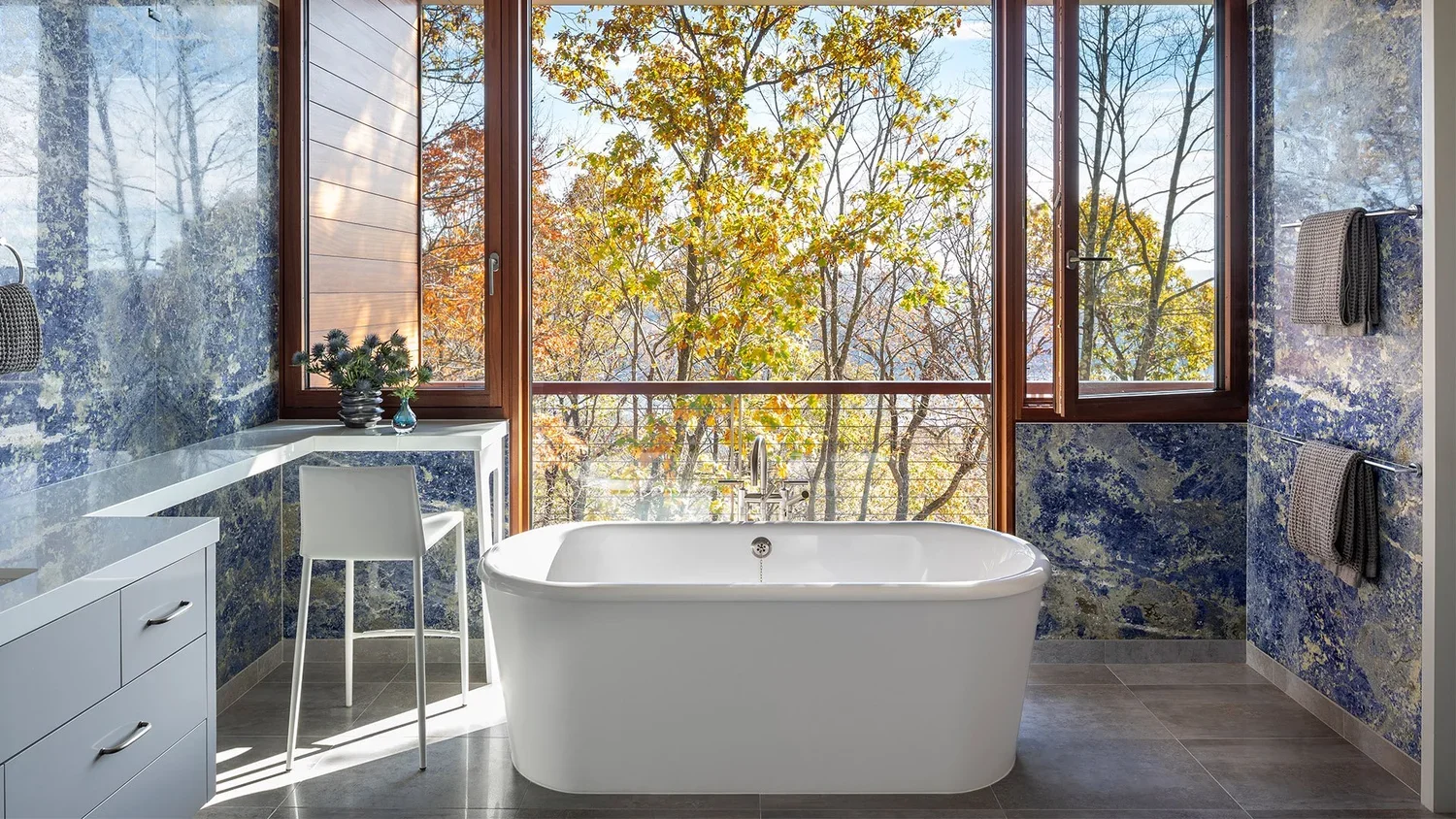 Modern bathroom with large window overlooking trees with autumn foliage, a white bathtub, a small white desk with a chair, potted plant, and blue and gray textured walls.